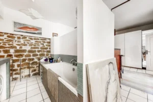 Salle de bain avec baignoire carrelée, mur en pierre apparente et espace fonctionnel.