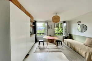Séjour lumineux avec vue arborée, sol en parquet blanc et coin repas dans un appartement à vendre à Paris 19, quartier Belleville.