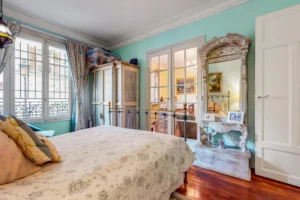 Chambre avec lit en fer forgé, armoire ancienne, miroir sculpté et lumière traversante.