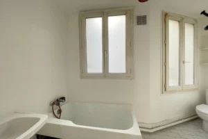 Salle de bains avec baignoire, fenêtres opaques et carrelage blanc, ambiance claire.
