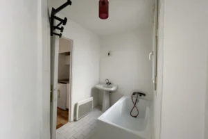 Salle de bains avec lavabo sur pied, vue sur la cuisine, murs blancs et lumière naturelle.