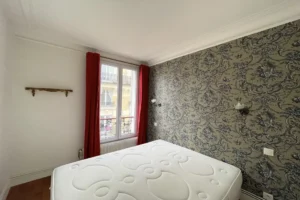 Chambre avec grande fenêtre, rideaux rouges et papier peint à motifs classiques dans un appartement parisien du 18ᵉ arrondissement, quartier Montmartre.