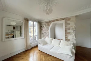 Séjour lumineux avec parquet, moulures au plafond et mur en pierre, vue sur les façades typiques du quartier des Abbesses à Paris 18ᵉ.