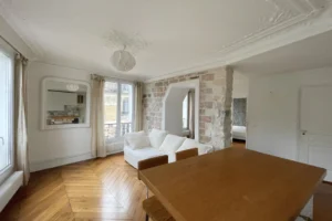 Pièce de vie élégante avec parquet ancien, moulures au plafond, mur en brique et lumière naturelle dans un appartement à Montmartre, Paris 18ᵉ.