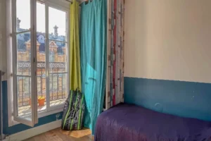 Chambre avec parquet ancien, lit double et porte-fenêtre ouvrant sur un balcon filant avec vue sur les toits parisiens, appartement situé rue du Faubourg Saint-Denis, secteur Gare du Nord à Paris 10.