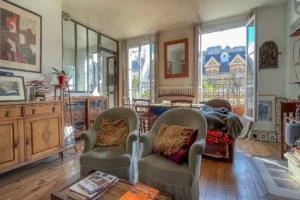 Salon lumineux avec parquet ancien, fauteuils et espace salle à manger ouvert, bénéficiant de larges fenêtres et d’un accès au balcon avec vue sur les toits parisiens, appartement situé rue du Faubourg Saint-Denis, secteur Gare du Nord à Paris 10.