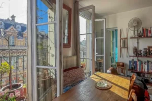 Espace repas baigné de lumière avec table en bois massif, ouvert sur un balcon filant grâce à de grandes portes-fenêtres, offrant une vue sur les façades parisiennes, appartement situé rue du Faubourg Saint-Denis, secteur Gare du Nord à Paris 10.