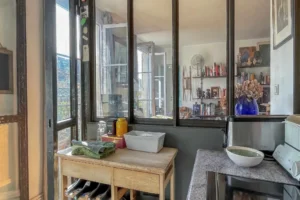 Cuisine séparée par une verrière de style atelier, avec plan de travail en pierre, rangements fonctionnels et ouverture sur la pièce de vie lumineuse, appartement situé rue du Faubourg Saint-Denis, secteur Gare du Nord à Paris 10.