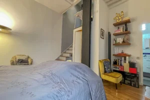 Chambre avec fauteuil jaune, bibliothèque murale et accès à la salle d’eau dans un appartement à vendre.