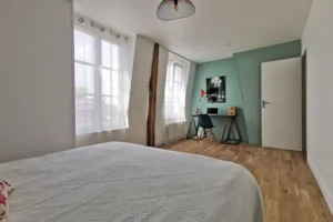 Chambre principale lumineuse avec deux fenêtres, coin bureau et parquet bois clair.