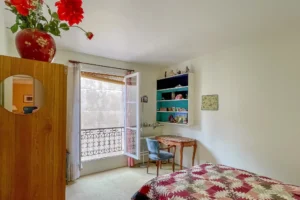 Chambre claire avec lit simple, bureau ancien, étagères murales et fenêtre sur cour.