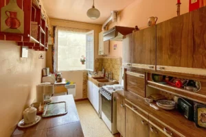 Cuisine indépendante à moderniser, équipée de meubles en bois et d’un évier sous fenêtre.