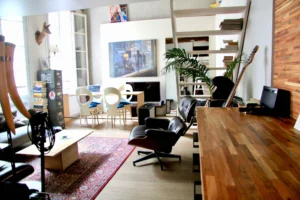 Vue générale du salon lumineux avec grande hauteur sous plafond, baies vitrées, fauteuils design, bibliothèque et coin repas dans un esprit arty et vintage – appartement à vendre Paris 4 Hôtel de Ville.