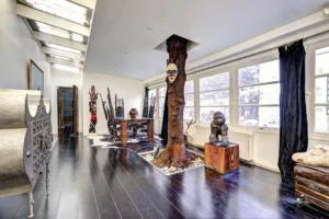 Loft à vendre Paris 17 – Pièce de vie traversante avec sol en bois sombre, grand arbre sculptural en son centre, mobilier ethnique et lumière naturelle traversante.