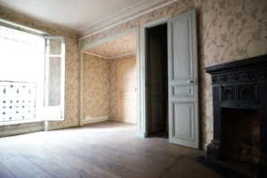 Pièce lumineuse avec parquet, cheminée ancienne en fonte, double exposition et papier peint à motif floral, dans un appartement à rénover à Belleville, Paris 19e.