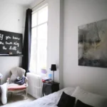Chambre cosy avec grande fenêtre, lit double, table de chevet noire et fauteuil ancien dans un loft à Paris 10ᵉ, rue du Faubourg Saint-Denis.