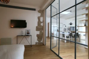 Chambre séparée par verrière, avec mur en pierre apparente, télé murale et vue dégagée sur l’espace de vie d’un loft à Paris 10.