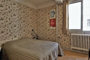 Chambre cosy d’un loft à Paris 10ᵉ avec papier peint fleuri, lit double, fenêtre sur cour et radiateur ancien.