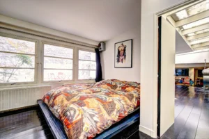 Chambre ouverte sur le séjour d’un loft à Paris 17 – Lit bas avec couverture graphique, grande baie vitrée sur cour et œuvre contemporaine au mur.