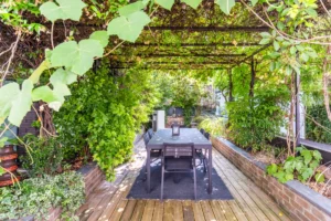 Salle à manger extérieure ombragée, sous une pergola végétalisée, avec table et chaises dans un cadre verdoyant.