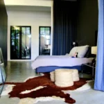 Chambre parentale élégante, avec rideaux bleu nuit, sol en béton ciré, tapis en peau et lumière naturelle filtrée par la verrière.