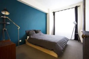 Chambre principale avec mur bleu intense, grand lit double et lumière naturelle filtrée par un voilage.