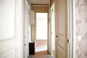 Entrée d’appartement avec couloir traversant, portes anciennes, boîtier technique apparent et parquet à restaurer, situé à Paris 19e, quartier Belleville.