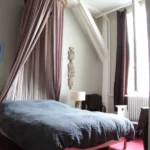 Chambre d’un loft à Paris 10 avec lit double, ciel de lit en tissu, sol bordeaux, mobilier en bois et lumière douce filtrée par de grandes fenêtres.