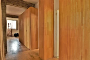 Couloir d’un loft à Paris 10ᵉ avec grands rangements en bois clair, parquet ancien et poutres apparentes en béton brut.