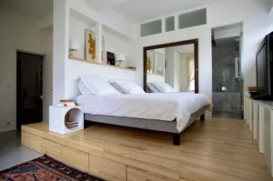 Suite parentale avec lit sur estrade en parquet clair, grand miroir mural et salle d’eau attenante, dans un appartement à vendre Paris 10ᵉ rue Martel.