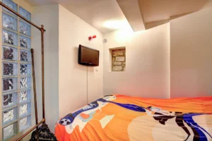 Chambre d’appoint dans le loft à Paris 17 – Mur en briques de verre, lit aux couleurs vives, télé murale et petite ouverture sur mur en pierre.
