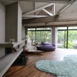 Salon d’un loft à Bagnolet avec cheminée en béton, canapé violet, parquet chaleureux et grandes baies vitrées donnant sur la végétation extérieure.