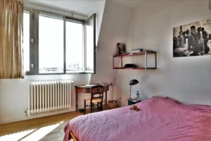 Chambre lumineuse dans un loft à Paris 10ᵉ avec lit double, bureau ancien, étagères et grande fenêtre avec vue dégagée sur les toits.