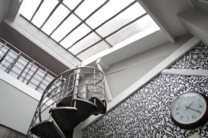 Escalier en spirale métallique sous une verrière de toit, avec mur à motif graphique et architecture industrielle dans un loft du 10ᵉ arrondissement de Paris.