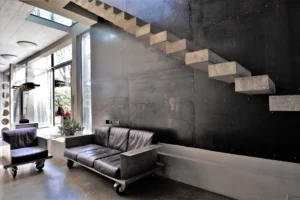 Escalier flottant en béton brut dans un loft industriel à Bagnolet, avec mobilier en métal et cuir sur roulettes et lumière naturelle abondante.