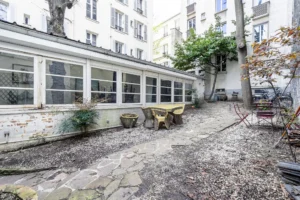 Extérieur d’un loft à Paris 17 – Cour privative arborée, mobilier en bois brut, chemin pavé et façades d’immeubles parisiens en arrière-plan.