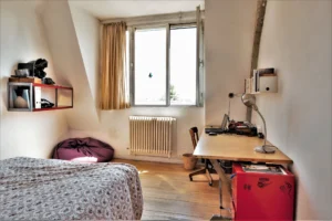 Chambre claire dans un loft à Paris 10ᵉ, avec lit double, grand bureau, étagères murales et pouf, sous une large fenêtre orientée sud.