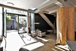 Entrée lumineuse d’un loft à Bagnolet, avec escalier en béton, poutres en bois brut, ouverture sur cour arborée et mobilier industriel sur roulettes.