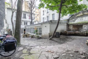 Cour arborée du loft à Paris 17 – Arbres en pleine terre, sol en pavés irréguliers, vélos, mur ancien en pierre et ambiance citadine paisible.