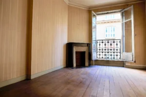 Vue d’une pièce à rénover avec cheminée ancienne, parquet massif et grande fenêtre ouverte sur cour intérieure, dans un appartement situé à Paris 19e, quartier Belleville.