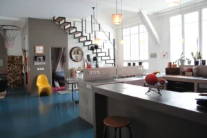 Grande cuisine ouverte au style industriel dans un loft parisien, avec îlot en béton, escalier graphique en métal noir, grandes baies vitrées et déco arty dans le quartier Gare de l’Est.