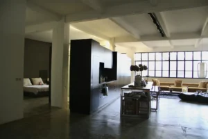 Cuisine ouverte noire au design minimaliste, attenante à l’espace nuit, avec grande verrière et table centrale dans un loft au style brut et fonctionnel.