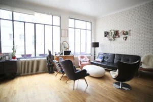Salon baigné de lumière naturelle avec parquet clair, mur en briques blanches, mobilier design et grandes fenêtres de type atelier dans un loft à Paris 10ᵉ.