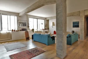 Grand salon de loft à Paris 10ᵉ avec parquet ancien, canapés colorés, tapis ethniques, poutres apparentes en béton brut et larges fenêtres rue de Chabrol.