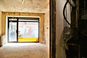 Espace intérieur brut du local commercial avec escalier en colimaçon vers l'étage, vitrine donnant sur rue commerçante.
