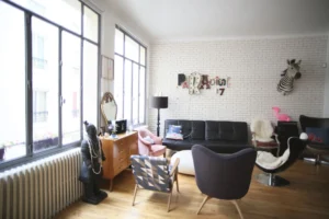 Coin salon lumineux avec canapé noir, fauteuils graphiques et décoration vintage dans un loft parisien au calme sur cour intérieure, quartier Gare du Nord.