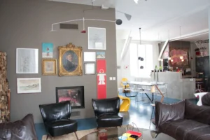 Salon d’un loft au style arty dans le 10e arrondissement de Paris, avec mur de cadres éclectiques, cheminée intégrée, fauteuils vintage et vue dégagée sur l’espace cuisine et salle à manger.