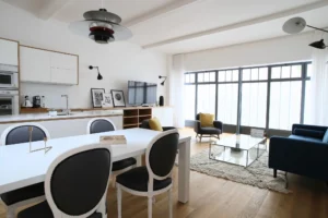 Pièce à vivre lumineuse dans un loft à Paris 10 avec verrière style atelier, coin salon cosy, fauteuils modernes et grande table à manger.