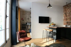 Cuisine ouverte dans un appartement à Montmartre Paris 18, avec mur en briques, îlot central en bois, fauteuil vintage et vue sur l’espace nuit en fond.