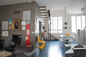 Salle à manger baignée de lumière dans un loft parisien, avec grande verrière, mobilier design, escalier métallique suspendu et murs décorés de cadres dans le quartier Faubourg Saint-Denis.